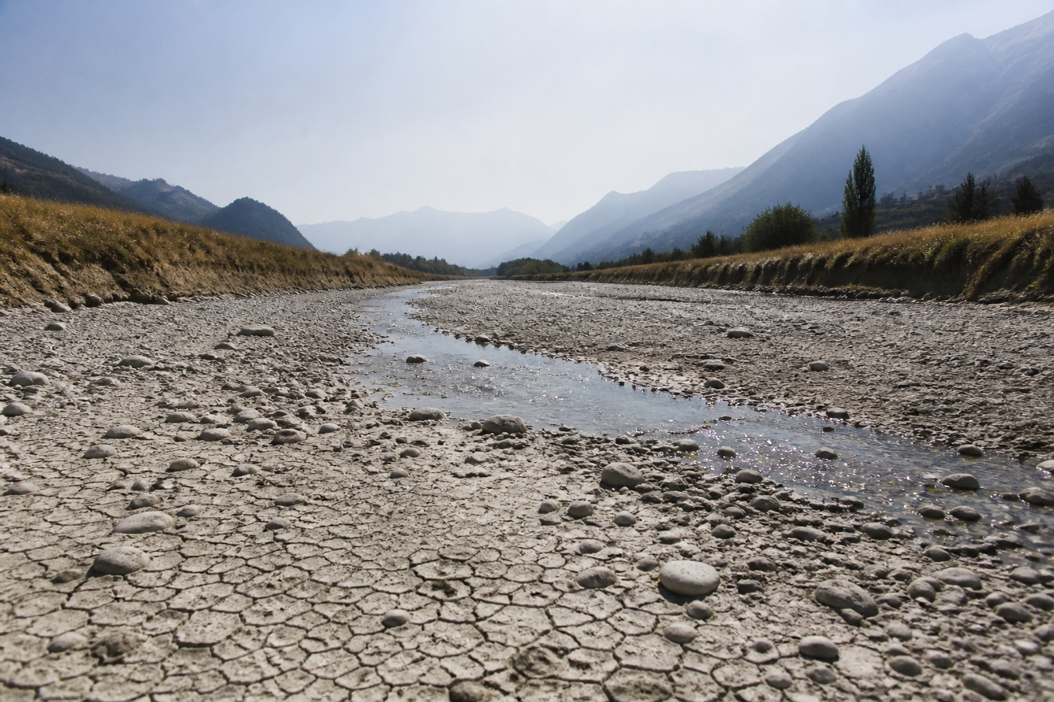 An almost dry riverbed of pale gravel with a thin thread of water below a Swiss valley