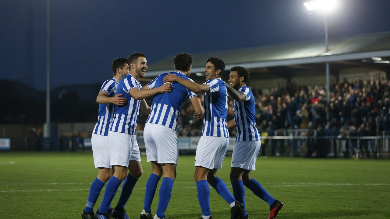 Footballers in blue and white shirts celebrating a goal in a small stadium under floodlights in the evening